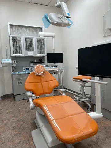 A treatment room with an orange dental chair and a large wall-mounted screen.